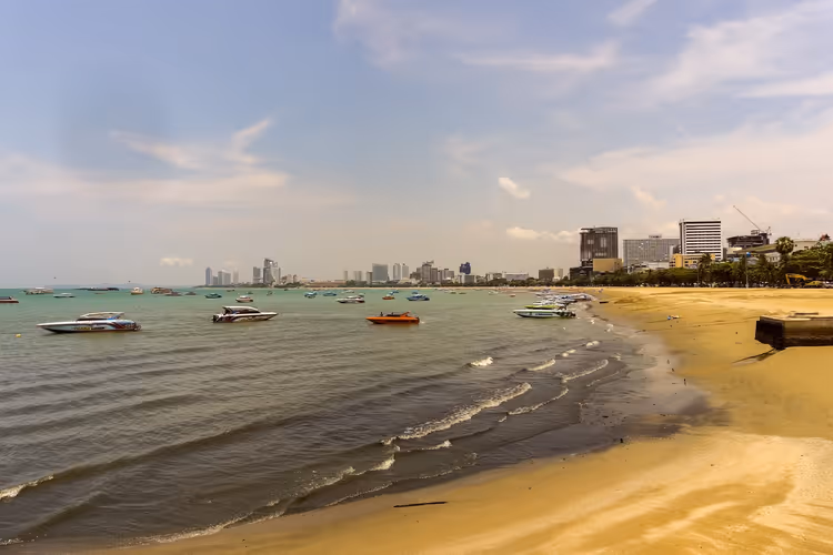 Schnellboote ankern vor einem Sandstrand in Pattaya, die Skyline der Stadt zieht sich unter dunstigem Himmel an der Bucht entlang
