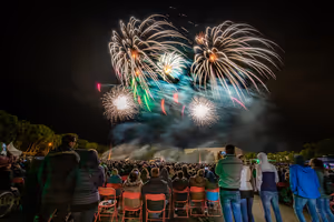 Menschenmenge beobachtet buntes Feuerwerk am Nachthimmel, einige Zuschauer sitzen auf roten Klappstühlen