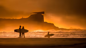 Silhouetten von Surfern mit Brettern am Strand bei Sonnenuntergang, Atlantikwellen und eine felsige Landzunge im orangefarbenen Dunst