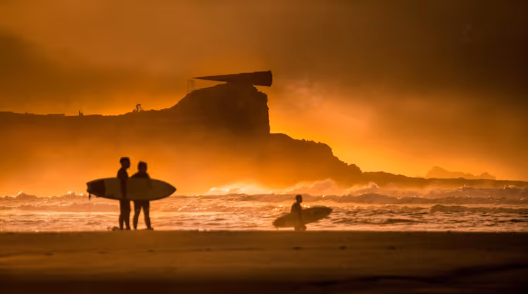 Silhouetten von Surfern mit Brettern am Strand bei Sonnenuntergang, Atlantikwellen und eine felsige Landzunge im orangefarbenen Dunst