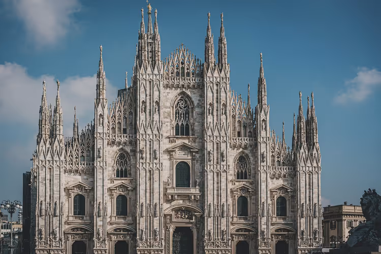 Gotische Fassade des Mailänder Doms aus weißem Marmor mit Reihen von Fialen und Statuen vor blauem Himmel mit leichten Wolken