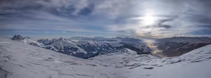 Panorama verschneiter Bergkämme unter einer dunstigen Sonne, tief unten ein beleuchtetes Tal