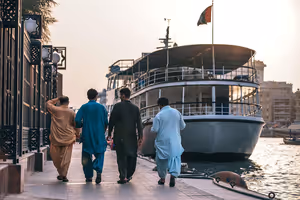 Vier Männer in traditionellen langen Gewändern gehen bei Sonnenuntergang am Kai des Dubai Creek an einem vertäuten Passagierboot vorbei