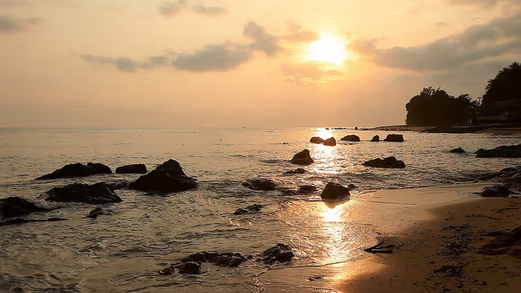 Goldener Sonnenuntergang über ruhigem Meer in Indonesien, mit dunklen Felsen im flachen Wasser und nassem Sandstrand