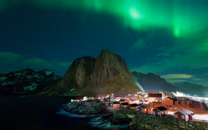 Grüne Polarlichter über roten Fischerhütten an einem felsigen Ufer unter einem steilen Berggipfel in Norwegen