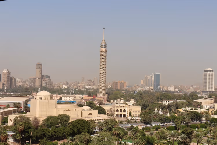 Kairotårnet med sitt gitterformede skaft over palmer, bygninger med kupler og en disig bysilhuett i Egypt