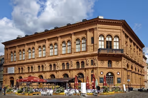 Reich verziertes terrakottafarbenes Gebäude im Palazzostil mit Bogenfenstern und Caféterrasse an einem gepflasterten Platz in Lettland