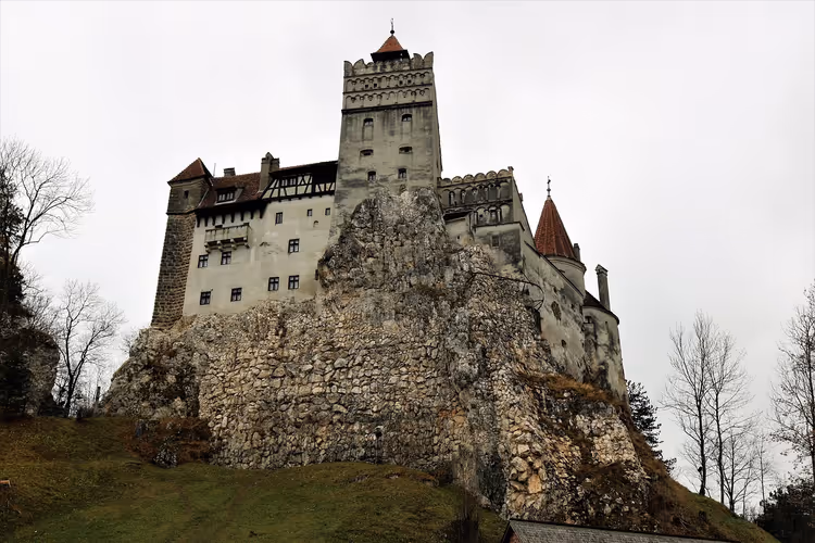 Schloss Bran in Rumänien auf einem Felsvorsprung, mit rot gedeckten Türmchen und kahlen Bäumen unter bedecktem Himmel
