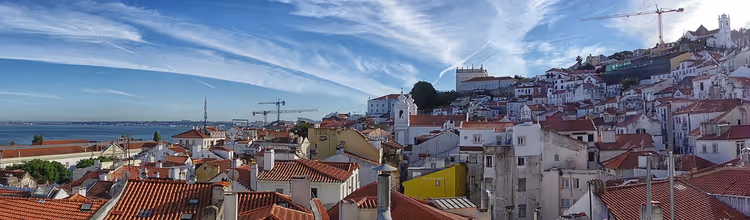 Panorama eines Hangs in Lissabon mit Terrakottadächern und weißen Häusern, links der Fluss, am Horizont Kräne