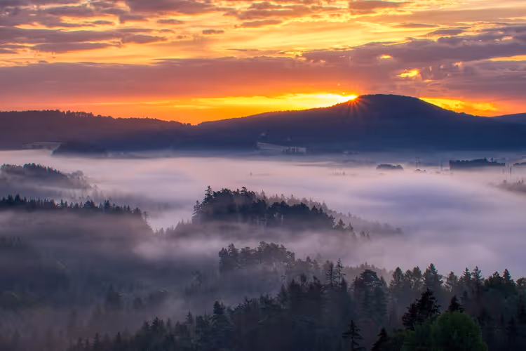 Sonnenaufgang über nebelgefüllten Tälern und bewaldeten Hügeln in der Böhmischen Schweiz, oranger Himmel hinter einem dunklen Kamm