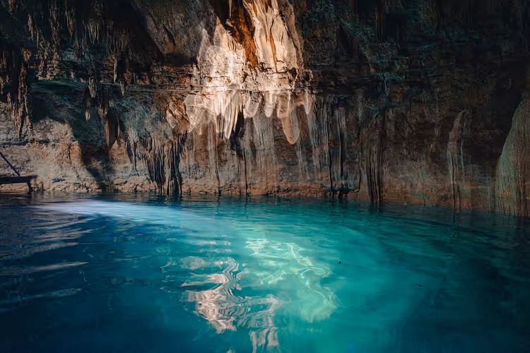Türkisfarbenes Wasser in einer Höhlen-Cenote, von einem Sonnenstrahl beleuchtet, an der Felswand hängen Stalaktiten