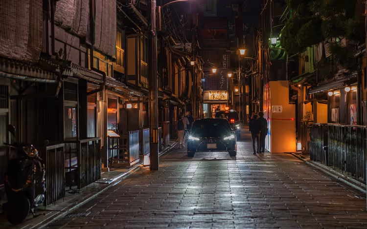 Schmale gepflasterte Gasse in Kyoto bei Nacht mit hölzernen Stadthäusern, leuchtenden Laternen und einem Auto mit Scheinwerferlicht