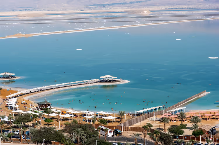 Türkisfarbene Bucht mit geschwungenem Steg, weißen Sonnenschirmen und Palmen an einem sandigen Hotelstrand, von oben gesehen