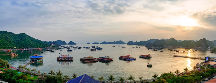 Schwimmende Häuser und Boote in einer Bucht in Vietnam, umgeben von Kalksteininseln unter dunstiger Abendsonne