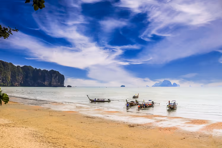 Longtailboote im seichten Wasser vor einem Sandstrand in Krabi, am Horizont Kalksteinfelsen und Inseln