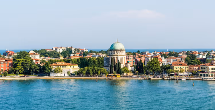 Chiesa dalla cupola verde e case dai tetti rossi lungo una riva alberata al di là della laguna turchese di Venezia