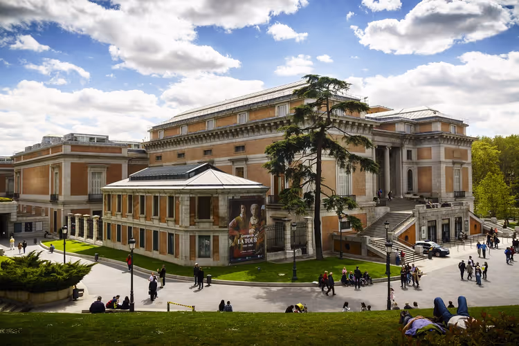 Prado-Museum in Madrid mit Fassade aus Backstein und Naturstein, einer hohen Zeder und Besuchern auf dem Rasen unter bewölktem Himmel