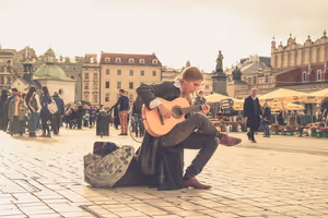 Straßenmusiker kniet mit Akustikgitarre auf einem gepflasterten Altstadtplatz, im Hintergrund Passanten