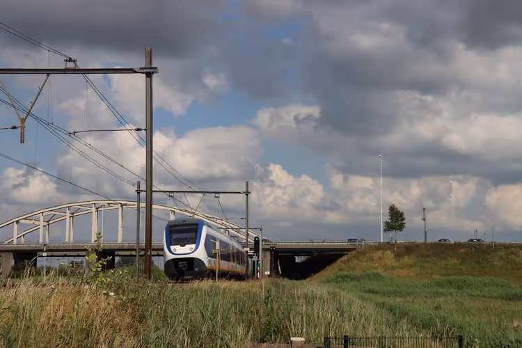 Blau-weißer Personenzug auf elektrifizierter Strecke an einer Bogenbrücke und einem grasbewachsenen Bahndamm unter bewölktem Himmel
