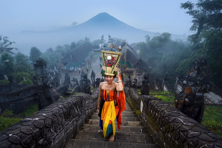 Frau in leuchtendem balinesischem Gewand trägt eine hohe Opfergabe auf dem Kopf eine Tempeltreppe hinauf, dahinter ein Vulkan im Dunst, Bali