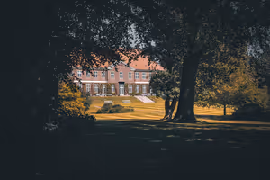 Herrenhaus aus rotem Backstein mit rotem Dach auf sonnigem, gestreift gemähtem Rasen, im Vordergrund von dunklen Bäumen gerahmt