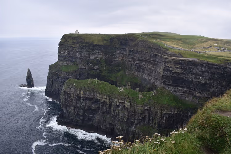 Geschichtete Meeresklippen mit grünem Gras und einem kleinen Turm, darunter eine Felsnadel im grauen Atlantik, Irland