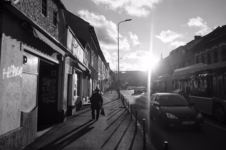 Straße in Brașov in Schwarz-Weiß, ein einzelner Fußgänger geht der tiefen Sonne entgegen, vorbei an alten Häusern und parkenden Autos