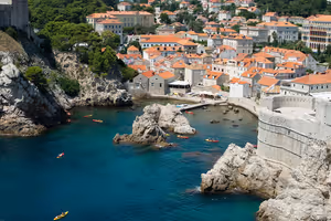 Kajaks in einer türkisfarbenen Bucht zwischen Felsklippen und Festungsmauern unterhalb der orangeroten Ziegeldächer von Dubrovnik