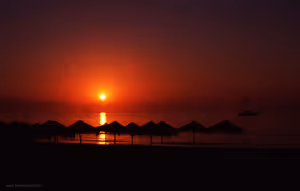 Silhouetten strohgedeckter Strandschirme vor einem tiefroten Sonnenaufgang über dem Meer, draußen ein Segelboot
