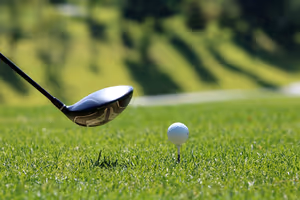 Kop van een golfdriver achter een witte bal op een tee, close-up op zonnig groen gras met een vage golfbaan erachter