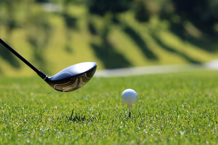 Kopf eines Golf-Drivers hinter einem weißen Ball auf dem Tee, Nahaufnahme auf sonnigem Grün, dahinter der unscharfe Platz