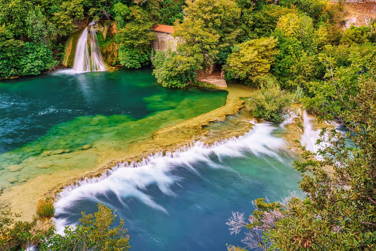 Smaragdgrüne Flussbecken und niedrige Kaskaden inmitten von dichtem grünem Wald in Kroatien, dahinter ein Wasserfall