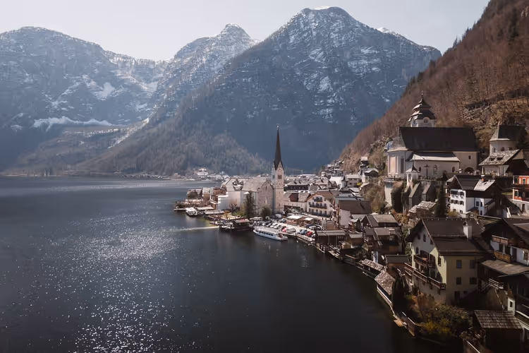 Dorf am See in den österreichischen Alpen mit spitzem Kirchturm, Booten auf dunklem Wasser und leicht verschneiten Gipfeln
