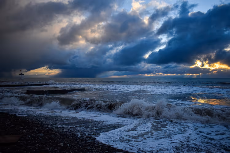 Stürmisches Meer mit schäumender Brandung an einem dunklen Kieselstrand unter schweren Wolken im Licht der tief stehenden Sonne