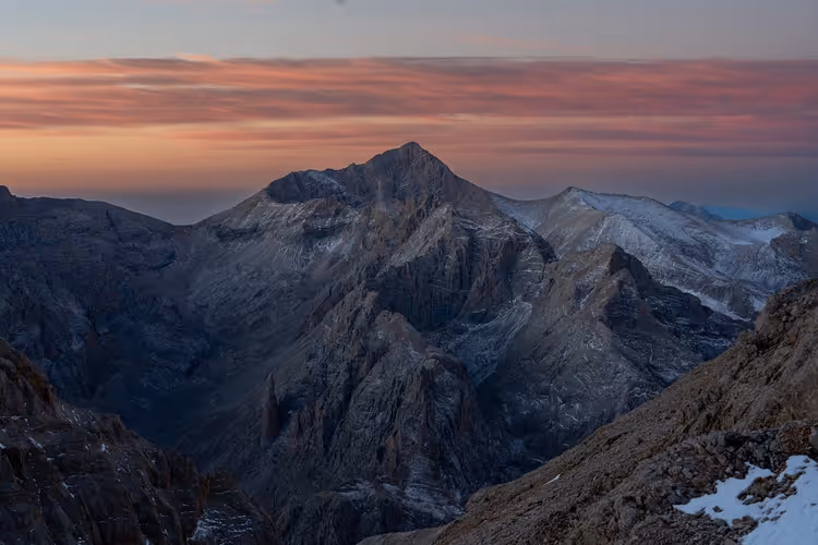 Schroffe, leicht verschneite Berggipfel in der Abenddämmerung, rosa und orange leuchtende Wolkenbänder über den dunklen Graten