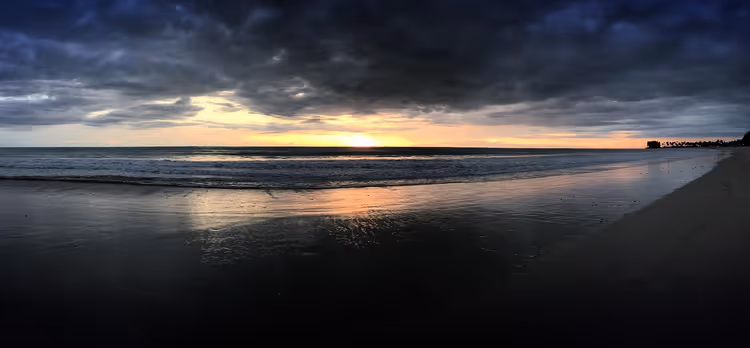 Strand bei Sonnenuntergang mit dunklen Wolken und Spiegelung im Sand