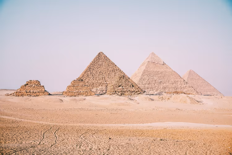Drei große Pyramiden und eine kleinere Stufenpyramide im hellen Wüstensand unter diesigem Himmel in Ägypten