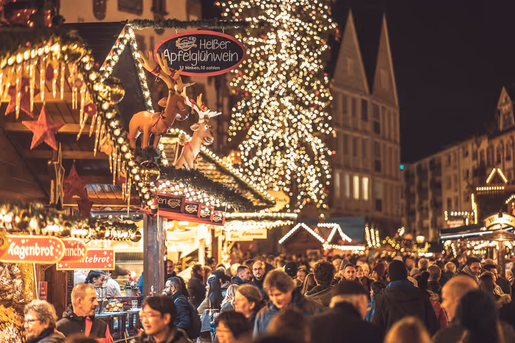 Voller Weihnachtsmarkt bei Nacht mit lichtergeschmückten Buden, Rentierfiguren auf einem Dach und einem leuchtenden Baum