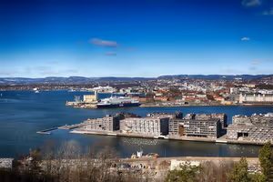 Uferfront von Oslo von einem Hügel aus, mit modernen Wohnblöcken auf einem Pier, einer Fähre im Fjord und fernen Schneehügeln