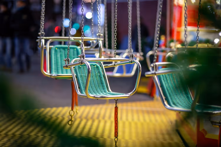 Leere grüne Sitze eines Kettenkarussells an Metallketten auf einem abendlichen Jahrmarkt mit unscharfen Lichtern