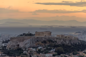 Akropolis von Athen mit dem Parthenon auf ihrem Felshügel bei Sonnenuntergang, dahinter ankernde Schiffe im dunstigen Golf