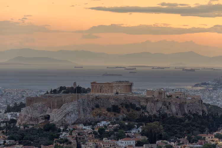 Akropolis von Athen mit dem Parthenon auf ihrem Felshügel bei Sonnenuntergang, dahinter ankernde Schiffe im dunstigen Golf
