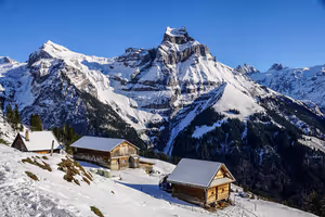 Holzchalets an einem verschneiten Hang mit Blick auf einen felsigen, schneebedeckten Alpengipfel unter klarem blauem Himmel