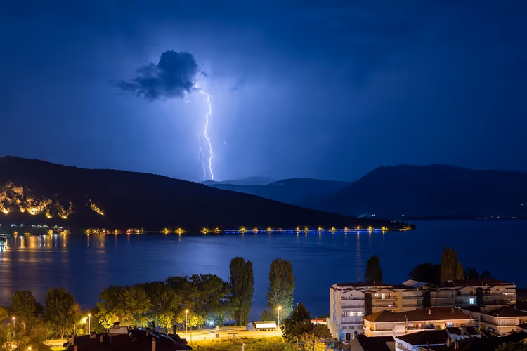 Blitz schlägt nachts aus einer dunklen Wolke über Hügeln ein, darunter ein beleuchtetes Ufer und ein Ort am See