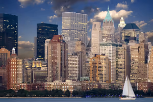 Dicht stehende Hochhäuser Manhattans in der Dämmerung vom Wasser aus, davor zieht ein weißes Segelboot vorbei, New York