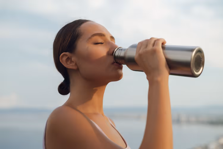 Mulher a beber água de uma garrafa térmica junto ao mar