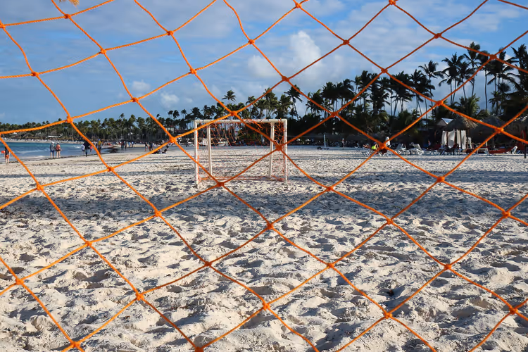 Weißer Sandstrand mit Fußballtor und Palmenreihen, durch ein orangefarbenes Netz gesehen, in der Dominikanischen Republik