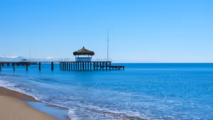 Holzsteg mit strohgedecktem Pavillon, der ins ruhige blaue Meer führt, neben einem Sandstrand unter klarem Himmel