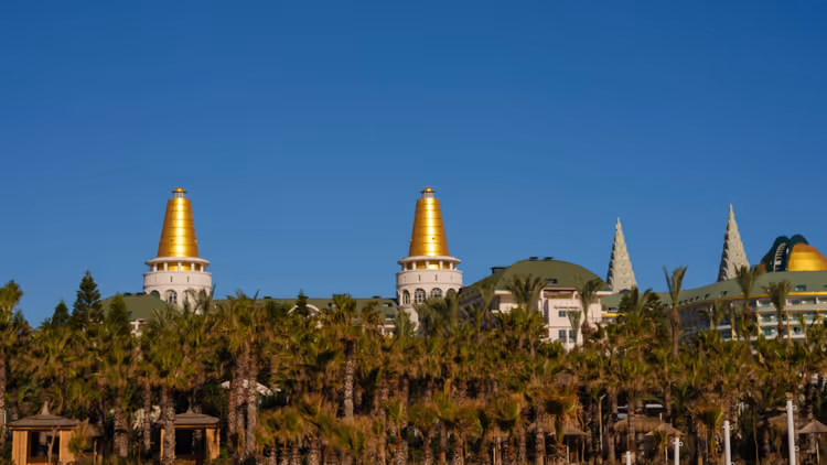 Türme eines Resorthotels mit goldenen Kuppeln über einer dichten Palmenreihe unter klarem blauem Himmel in Belek
