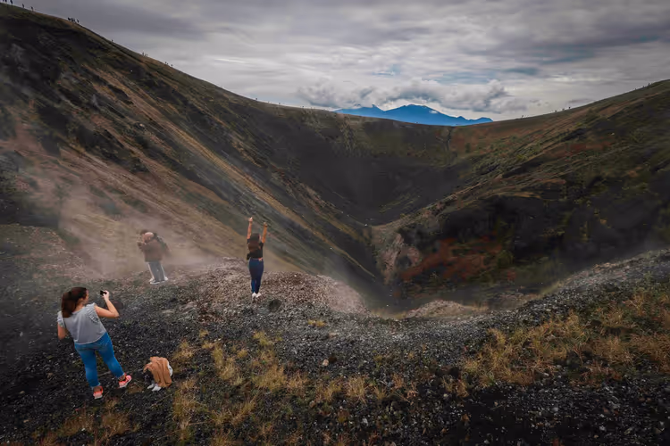 Wanderer auf dem dunklen Schotterrand eines Vulkankraters, eine Frau hebt die Arme, jemand fotografiert, bewölkter Himmel